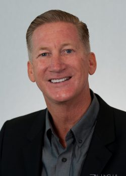 A middle-aged man in a suit jacket and collared shirt smiles at the camera against a plain background.
