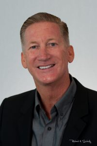 A middle-aged man in a suit jacket and collared shirt smiles at the camera against a plain background.
