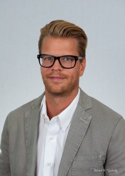 A man with blond hair, glasses, and a beard wears a light gray blazer over a white shirt, facing the camera.