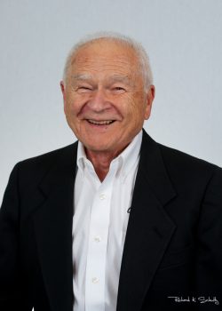 An older man in a black suit jacket and white shirt smiles at the camera against a plain light background.