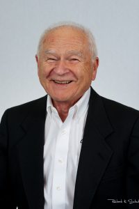 An older man in a black suit jacket and white shirt smiles at the camera against a plain light background.