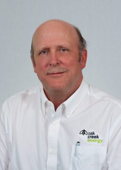 A middle-aged man in a white Oak Creek Energy shirt poses for a professional portrait against a light gray background.