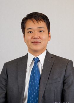 A man in a gray suit, white shirt, and blue tie poses against a plain light background.