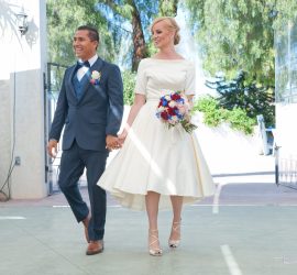 A bride in a white dress and a groom in a blue suit walk hand in hand outdoors on their wedding day.