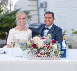 Bride and groom sit at a decorated table with champagne and flowers, sign reads "The Calzadas.