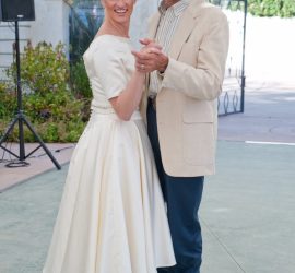 A woman in a white dress and a man in a beige jacket and cowboy hat dance together outdoors.