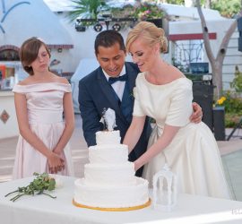 A bride and groom cut a white, three-tier wedding cake while a bridesmaid stands beside them outdoors.