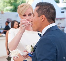 A bride in a white dress feeds cake to a groom in a suit during an outdoor wedding reception.