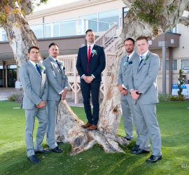 Five men in suits pose for a photo outdoors by a large tree, with one man standing on the tree roots.