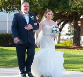 A bride in a white dress walks down an outdoor path arm-in-arm with a man in a suit, both facing forward.