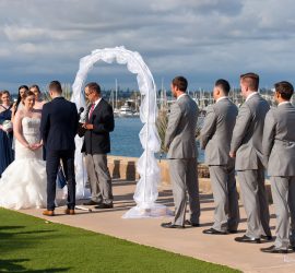 A bride and groom stand under a white arch by the water, surrounded by their wedding party and an officiant.