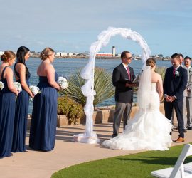 A bride and groom stand under an arch by the water during an outdoor wedding ceremony with bridal party present.