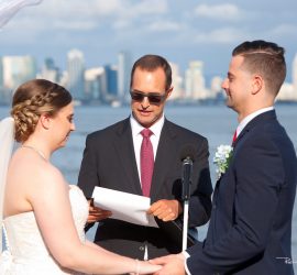 A bride and groom hold hands during an outdoor wedding ceremony with an officiant, city skyline in the background.
