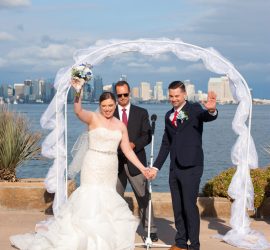 A bride and groom stand under a white arch, holding hands and waving, with a city skyline and water in the background.
