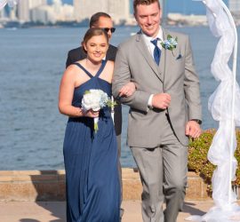 A bridesmaid in a navy dress and a groomsman in a gray suit walk arm-in-arm outdoors by the water under a white arch.