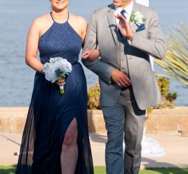 A woman in a navy dress and a man in a gray suit walk arm-in-arm outdoors by the water, holding bouquets.