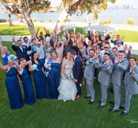 A wedding group photo outdoors shows the bride and groom kissing, surrounded by cheering guests in formal attire.