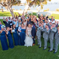 A wedding group photo outdoors shows the bride and groom kissing, surrounded by cheering guests in formal attire.