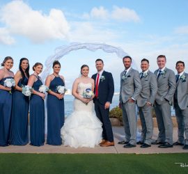 A bride and groom stand with four bridesmaids in blue dresses and four groomsmen in gray suits by the water.