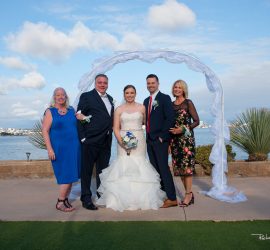 Five people pose for a wedding photo outdoors by the water, standing under a white arch with a scenic background.