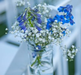 A mason jar with blue and white flowers is attached to a white chair, with more chairs blurred in the background.