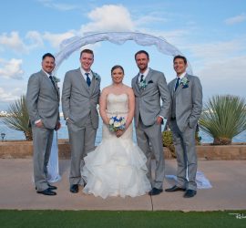A bride in a white gown stands with four men in matching grey suits under an arch by the water on a sunny day.