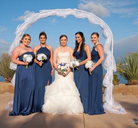 Five women, including a bride in white and four bridesmaids in navy blue dresses, pose outdoors under a white arch by the water.