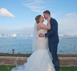Bride and groom embrace and kiss outdoors by the water, with a bouquet on the grass and city skyline in the background.