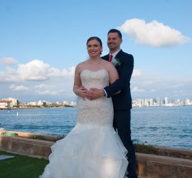 A bride and groom stand together outdoors near the water, with a city skyline and clouds in the background.