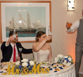 Three people at a wedding reception raise drinks in a toast, with "Mr & Mrs" sign and ship painting in the background.