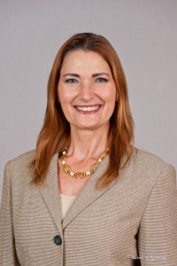 Woman with long brown hair wearing a beige blazer and gold necklace, smiling at the camera against a plain background.