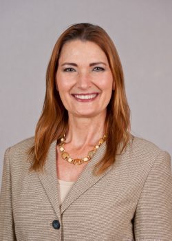 Woman with long brown hair wearing a beige blazer and gold necklace, smiling at the camera against a plain background.