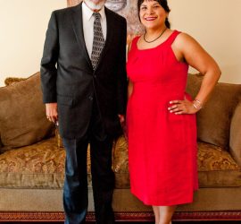 A man in a black suit and a woman in a red dress stand side by side in a living room, smiling at the camera.