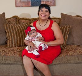Woman in a red dress sits on a couch, holding a baby in a matching red and white outfit with a bow headband.