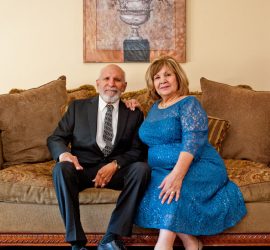 An older man in a suit and an older woman in a blue dress sit together on a couch in a living room.