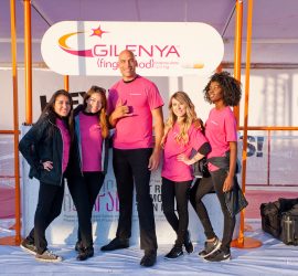 Five people in matching pink shirts stand posing in front of a Gilenya promotional sign at an outdoor event.