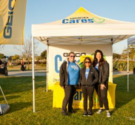 Three women stand in front of a GEICO Cares tent and table at an outdoor event on a sunny day.