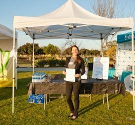 A woman stands and smiles in front of an outdoor information booth, holding a flyer, with event tents in the background.