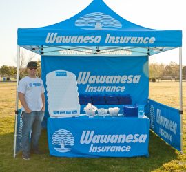 A man stands beside a Wawanesa Insurance booth with promotional materials and a plinko game at an outdoor event.