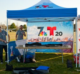Two people set up a Telemundo 20 and NBC 7 canopy tent at an outdoor event on a grassy field.