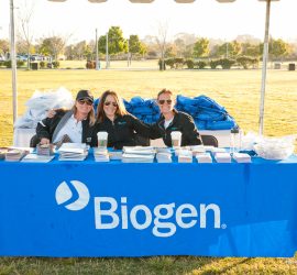 Three women sit behind a Biogen-branded table with pamphlets and bags at an outdoor event.