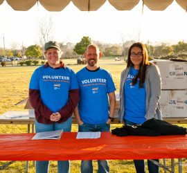 Three volunteers stand behind a table outdoors, wearing blue volunteer shirts and smiling at the camera.