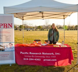 A woman stands behind a Pacific Research Network booth at an outdoor event, with brochures and promotional items on the table.