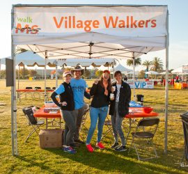 Four people stand and smile under a "Village Walkers" tent at a Walk MS event in a grassy outdoor area.