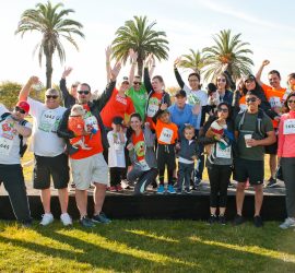 Group of people, including children and dogs, pose and celebrate on a stage outdoors with palm trees in the background.