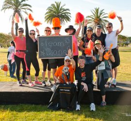 A group of people outdoors holds a "Team Diane" sign, wearing athletic clothes and orange accessories, posing and smiling.
