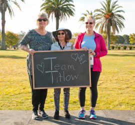 Three women stand outdoors holding a chalkboard sign that reads "Team Titlow" with palm trees in the background.