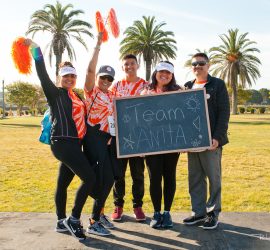 Five people in matching shirts stand outdoors holding a chalkboard that says "Team Anita," with palm trees in the background.
