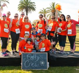A group in matching orange shirts poses outdoors with a "Cathy's Crew" sign, some holding orange pom-poms.
