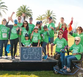 A group of people in green "Team Otis" shirts pose outdoors with a chalkboard sign, some raising their hands and cheering.